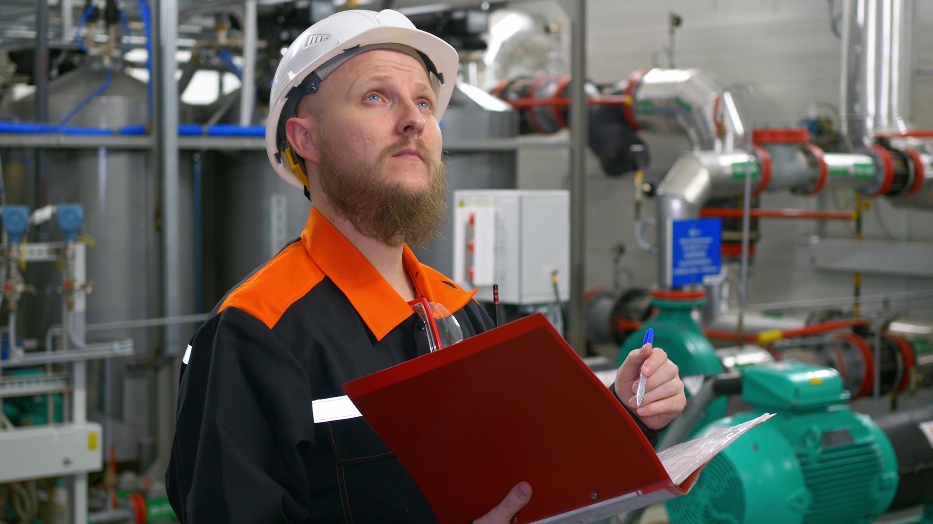 A bearded man, a mechanical engineer in a white helmet, makes an audit of equipment in an industrial workshop, in his hands is a pen with a folder in which there is a list of equipment.